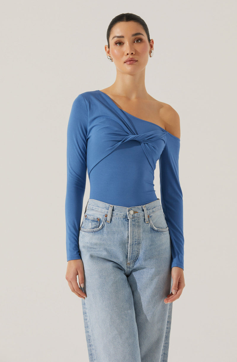 A woman is wearing a fitted, long-sleeved, royal blue top with a unique, twist knot detail on the front and an off-the-shoulder design on her left side. She is also wearing high-waisted, light-wash jeans with a relaxed fit.