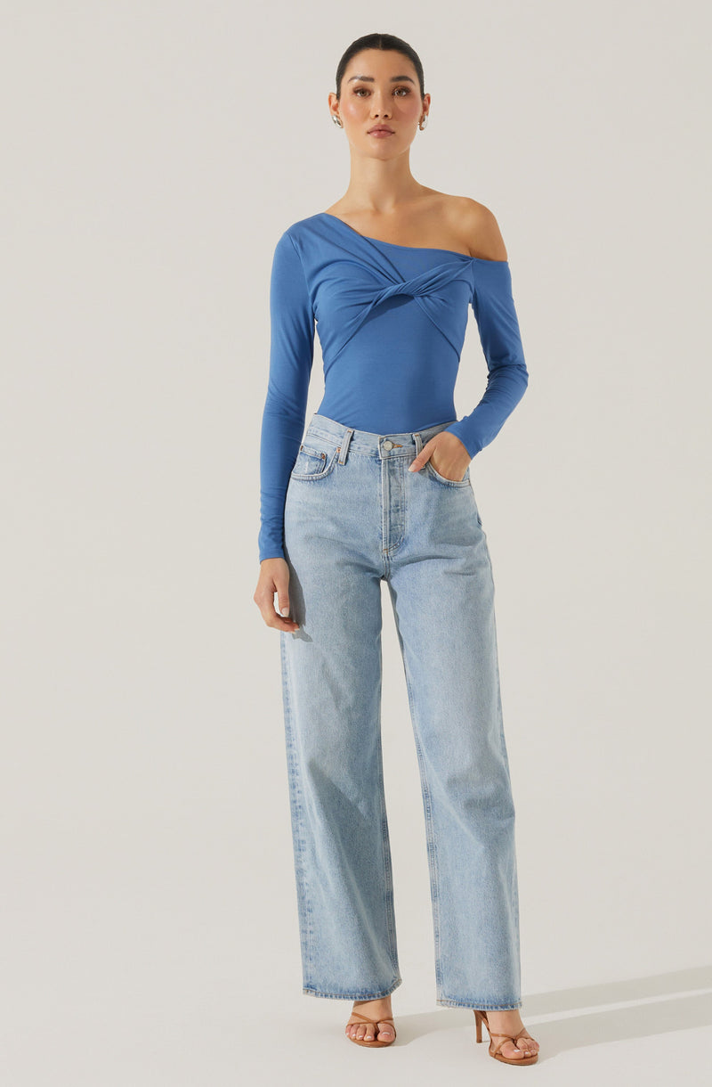 A woman is wearing a fitted, long-sleeved, one-shoulder blue top with gathered and twisted detailing at the bust. She pairs it with light-wash, high-waisted, wide-leg jeans and beige high-heeled sandals.