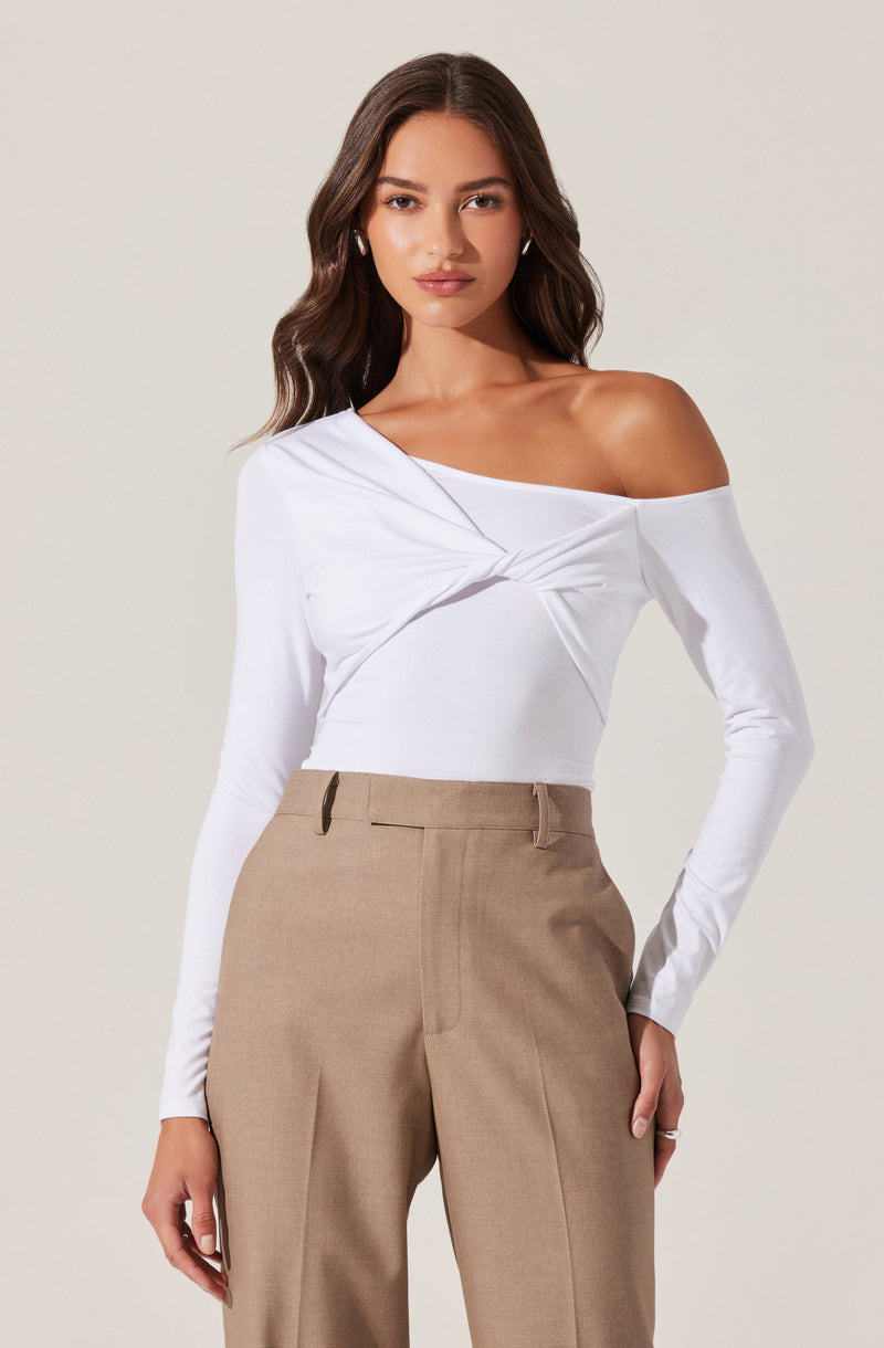 A woman is wearing a white, off-the-shoulder long-sleeve top with a twisted knot detail on the front. She pairs it with high-waisted, beige tailored trousers.