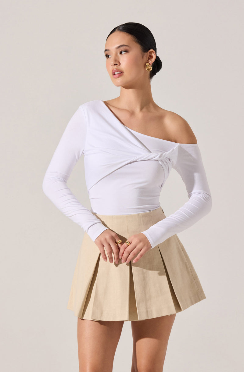 A woman is wearing a white, long-sleeved, off-the-shoulder top with a gathered detail across the front. She pairs it with a beige, pleated, high-waisted mini skirt.