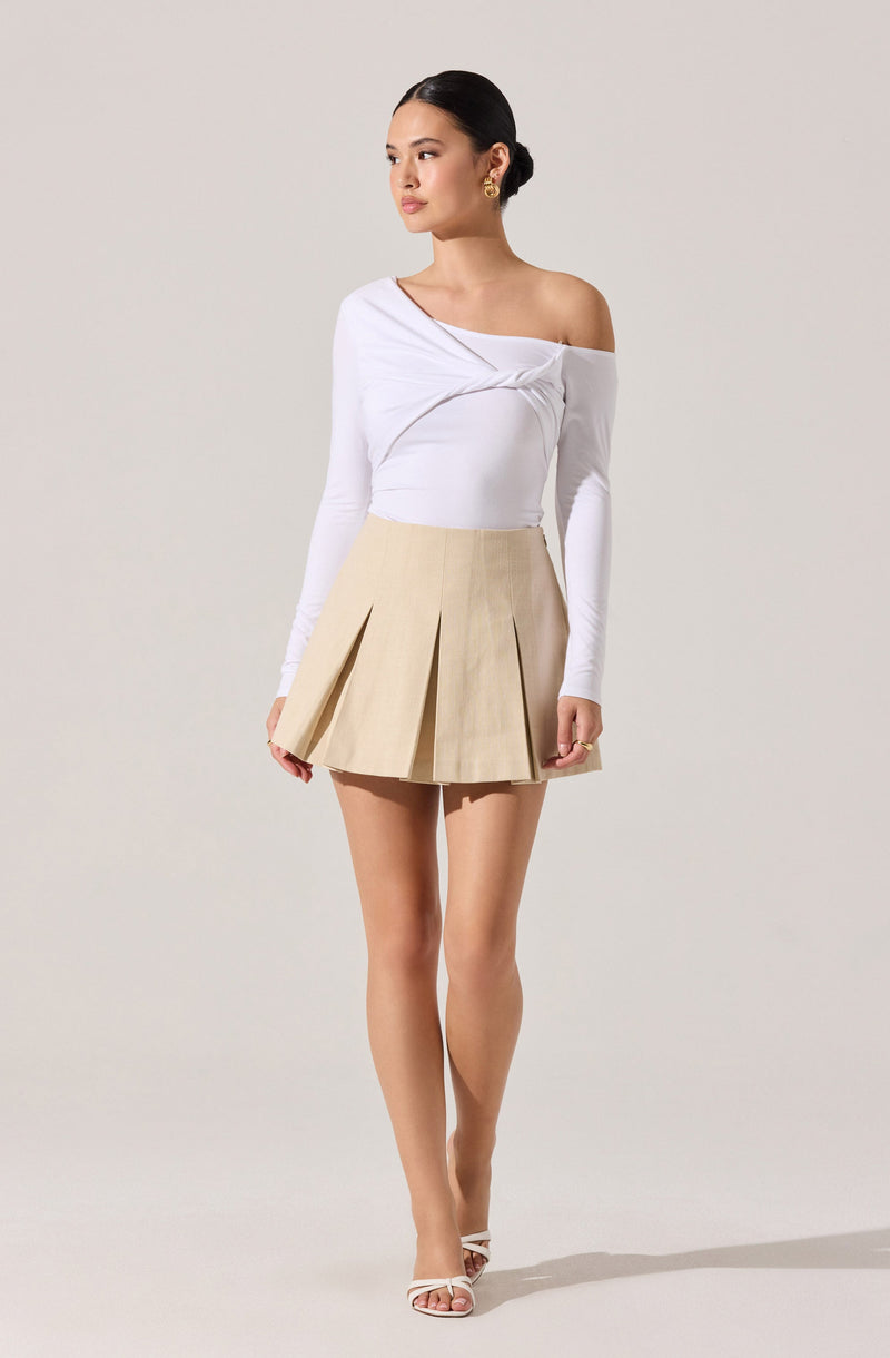 The woman is wearing a white, long-sleeved, off-the-shoulder top with a draped, twisted design across the chest. She is also wearing a high-waisted, beige pleated mini skirt and white strappy high-heeled sandals. She has gold earrings and her hair is styled in a sleek, low bun.