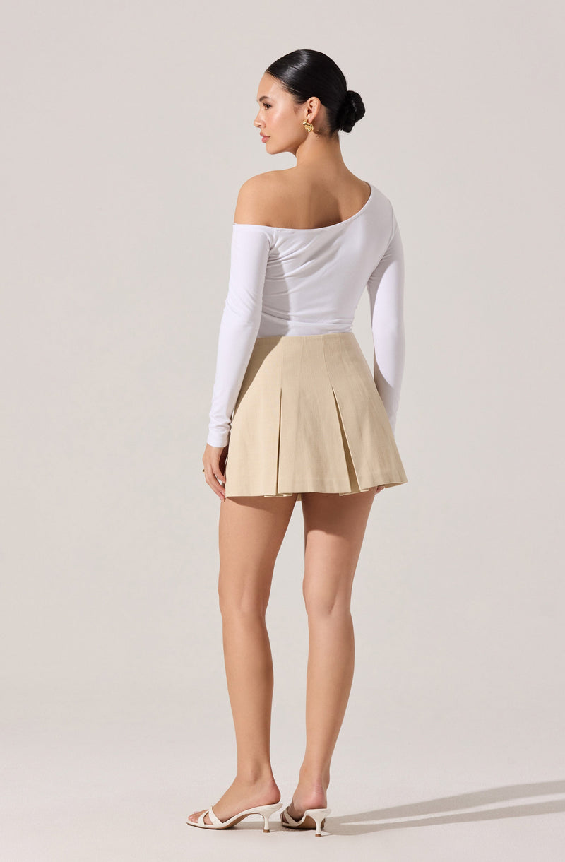 A woman is wearing an off-the-shoulder white long-sleeve top that is fitted and has a slight sheen, paired with a high-waisted beige pleated mini skirt with prominent front folds. She is also wearing white open-toe heeled sandals.