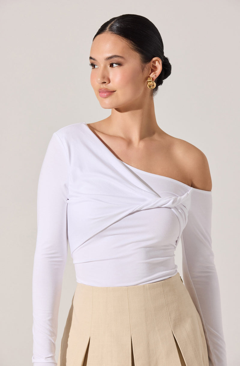 A woman is wearing a white, long-sleeve, one-shoulder top with a twist detail across the chest, paired with high-waisted beige pleated trousers. She has her hair neatly styled in a low bun and is accessorizing with gold hoop earrings.