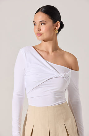 A woman is wearing a white, long-sleeve, one-shoulder top with a twist detail across the chest, paired with high-waisted beige pleated trousers. She has her hair neatly styled in a low bun and is accessorizing with gold hoop earrings.
