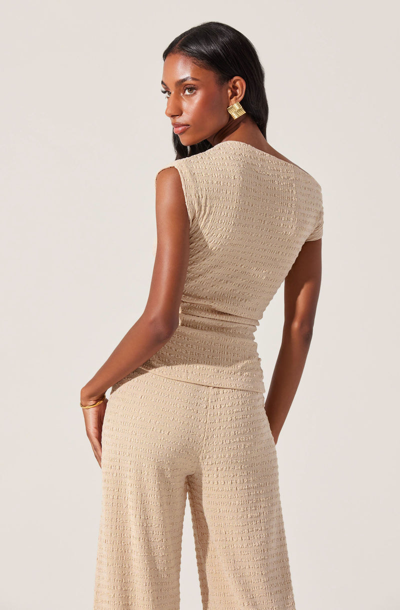 A woman is wearing a matching beige sleeveless top and wide-leg pants set. The fabric has a textured, slightly ribbed pattern. The top has a high, round neckline and armholes that slightly extend over the shoulders. The pants have a relaxed fit, with a straight waistband and no visible belt. The overall look is elegant and monochromatic.