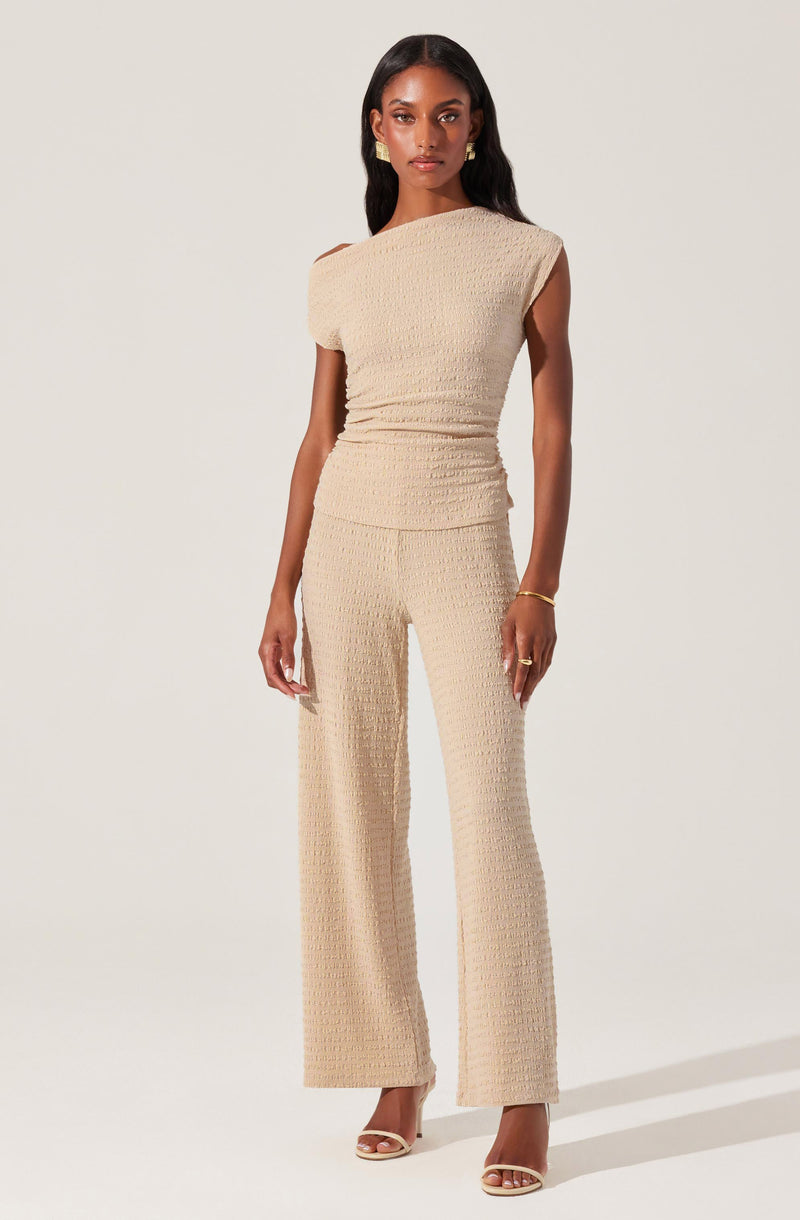 A woman is wearing a matching beige textured knit sleeveless top and wide-leg pants. The top has a boat neckline and slightly caped shoulders, while the pants are high-waisted and flow loosely down to her ankles. She is accessorized with gold earrings, a bracelet, and rings, and is wearing open-toe heels.