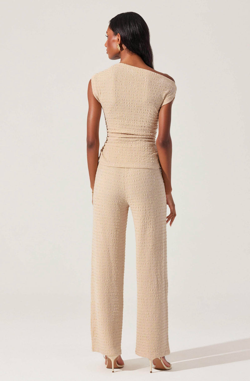 A woman is wearing a matching beige outfit consisting of a sleeveless, textured top with a round neckline and wide-leg pants with a similar textured pattern. She is also wearing high-heeled shoes.