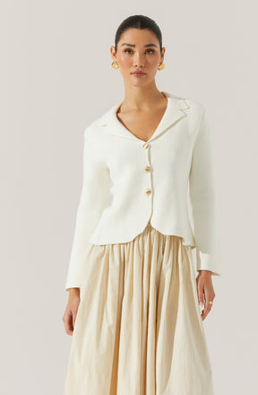 A woman is wearing an elegant white blazer with a notched lapel collar and large pearl-like buttons down the front. The blazer has a slightly textured fabric and a tailored fit. She is also wearing a flowing, high-waisted, beige skirt with gathered fabric, creating a voluminous and soft silhouette. She accessorized with gold earrings and a ring.