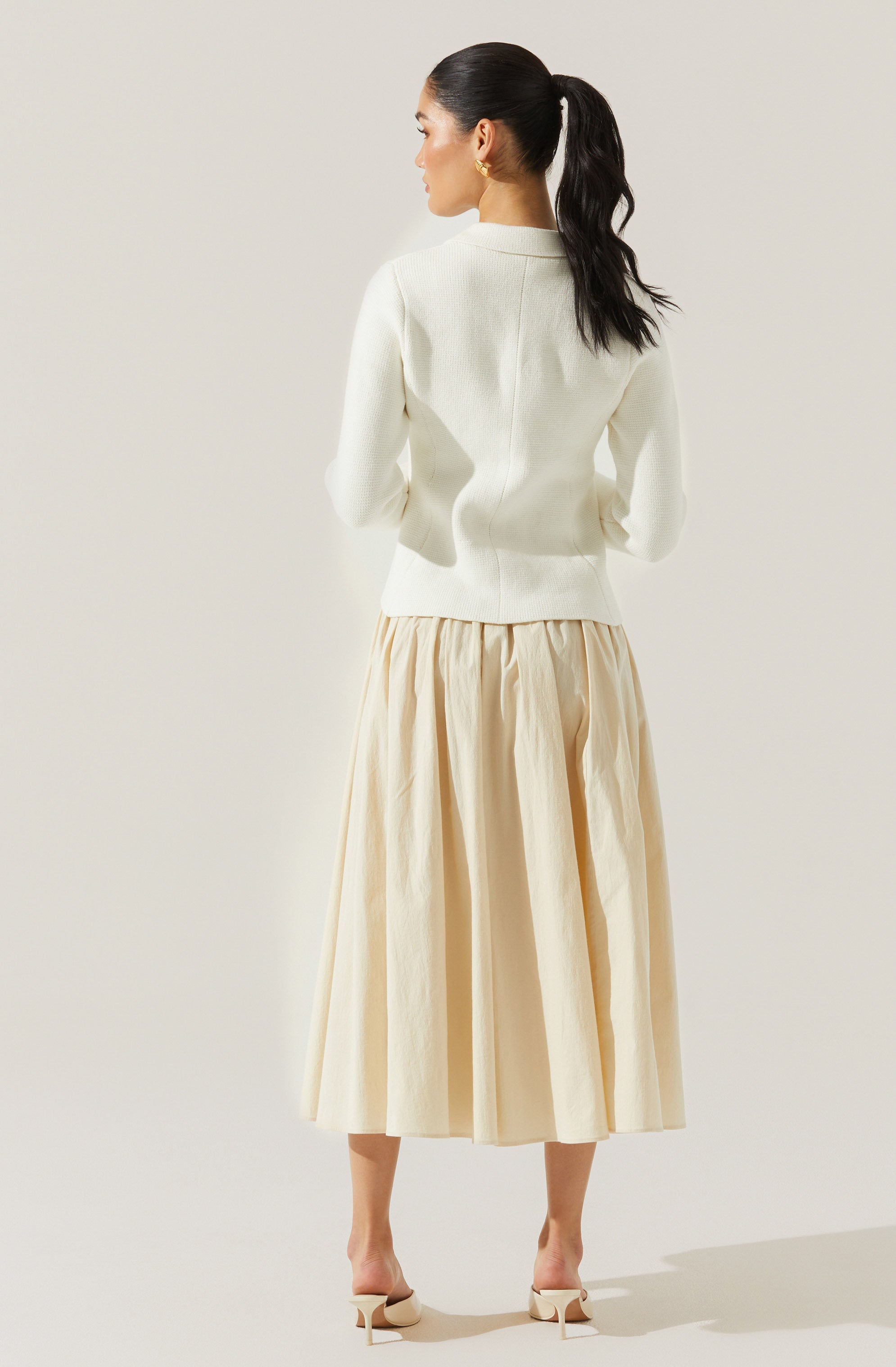 The woman is wearing a white long-sleeve sweater with a high neckline, paired with a beige, full-length, pleated skirt. She has on light-colored high-heeled shoes and gold hoop earrings. Her hair is styled in a high ponytail.