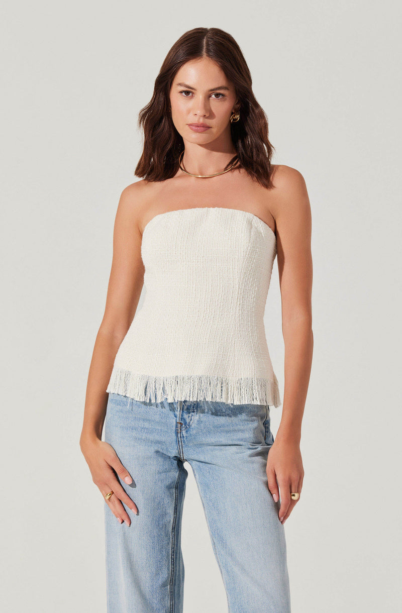 A woman is wearing a strapless, off-white, textured top with a frayed hem, paired with light-wash jeans.