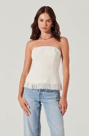A woman is wearing a strapless, off-white, textured top with a frayed hem, paired with light-wash jeans.