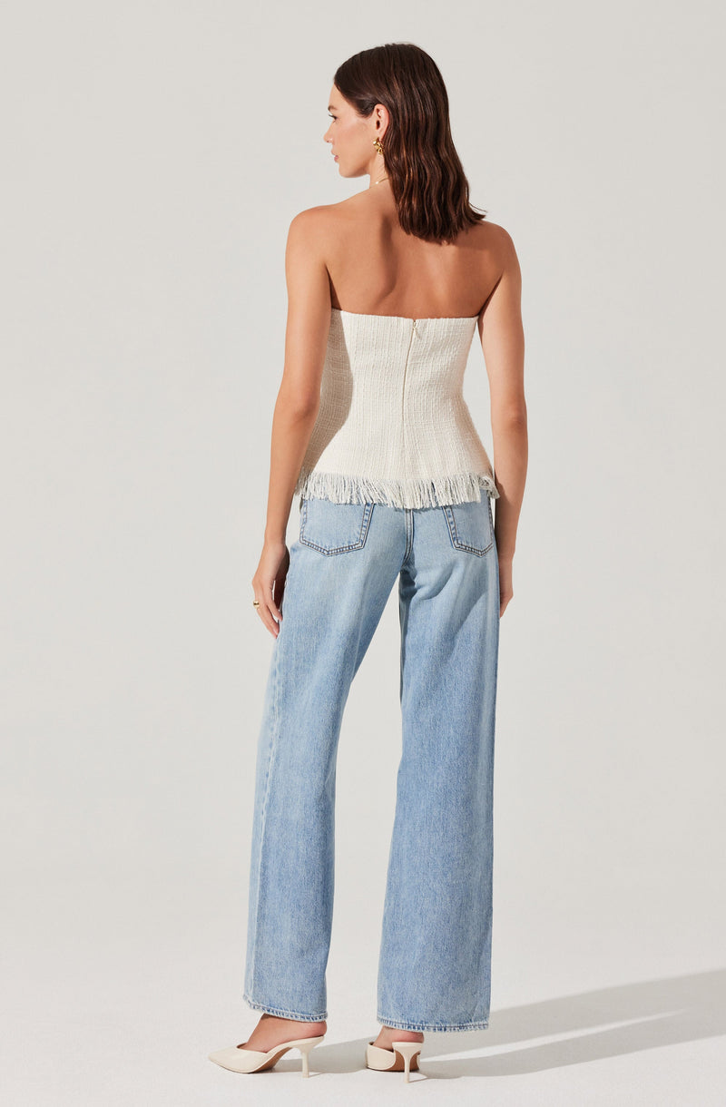 A woman is wearing a strapless, cream-colored, textured top with fringe detail at the hem, paired with light blue, high-waisted, wide-leg jeans. She is also wearing off-white, pointed-toe heels with a block heel.
