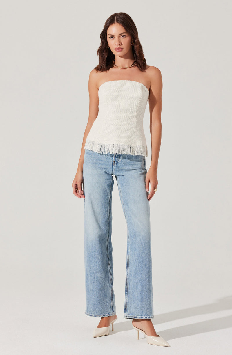 A young woman is wearing a strapless, off-white top made of textured fabric with a fringe hem at the bottom. She is also dressed in light-wash, high-waisted, wide-leg jeans that reach down to her heels. She pairs the outfit with white pointed-toe high heels.
