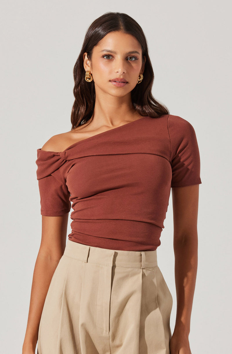A woman is wearing a fitted, off-the-shoulder top in a warm terracotta color with short sleeves and a ruched detail across the chest. She pairs it with high-waisted, beige pleated trousers.
