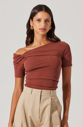 A woman is wearing a fitted, off-the-shoulder top in a warm terracotta color with short sleeves and a ruched detail across the chest. She pairs it with high-waisted, beige pleated trousers.