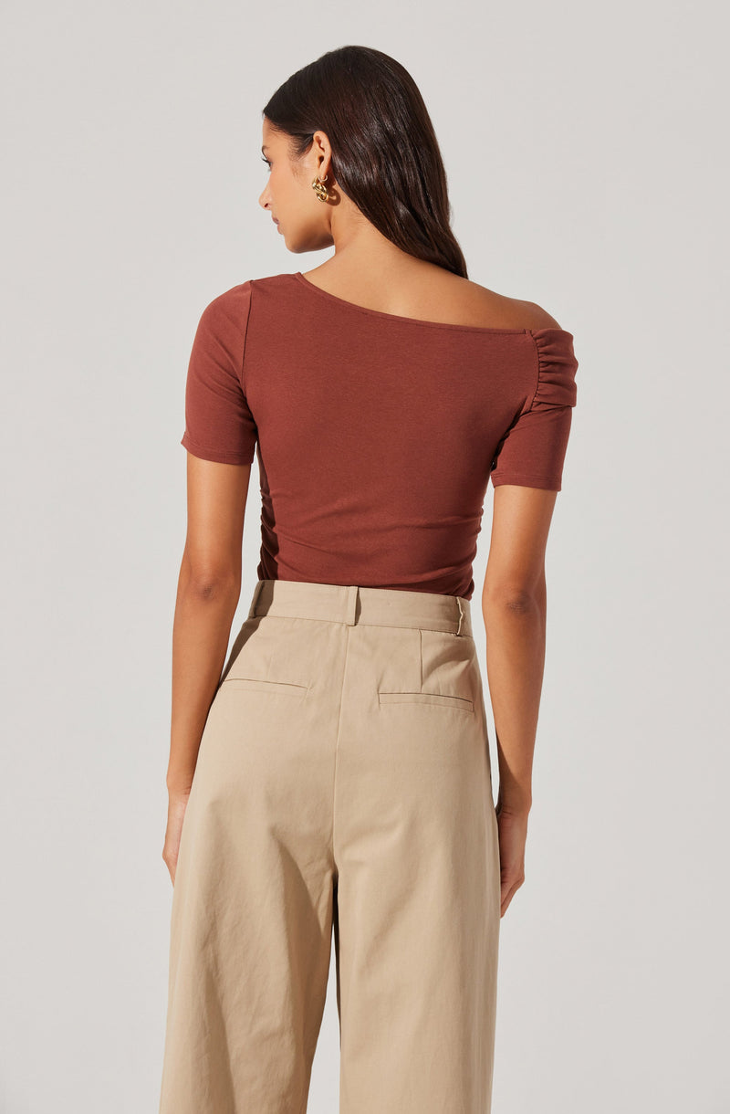 A woman is wearing a fitted, short-sleeved, rust-colored top with a wide neckline that drapes over her shoulders and ruched detailing on the left shoulder. She pairs it with high-waisted, beige, wide-leg trousers that have a tailored fit.