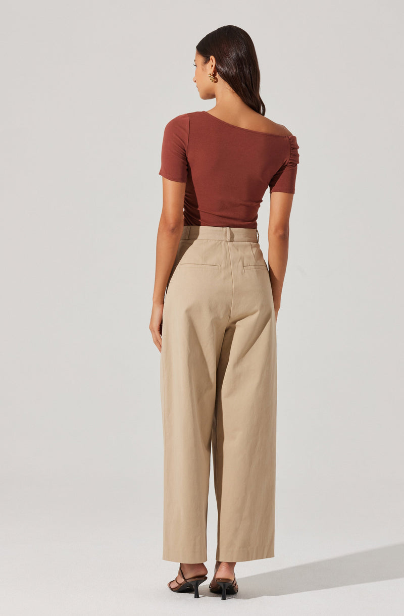 A woman is wearing a fitted, short-sleeved top in a warm, rusty brown color with a wide neckline that slouches slightly off one shoulder. She pairs it with high-waisted, wide-leg beige trousers that have a relaxed fit and a classic waistband with belt loops. The outfit is completed with black high-heeled sandals.