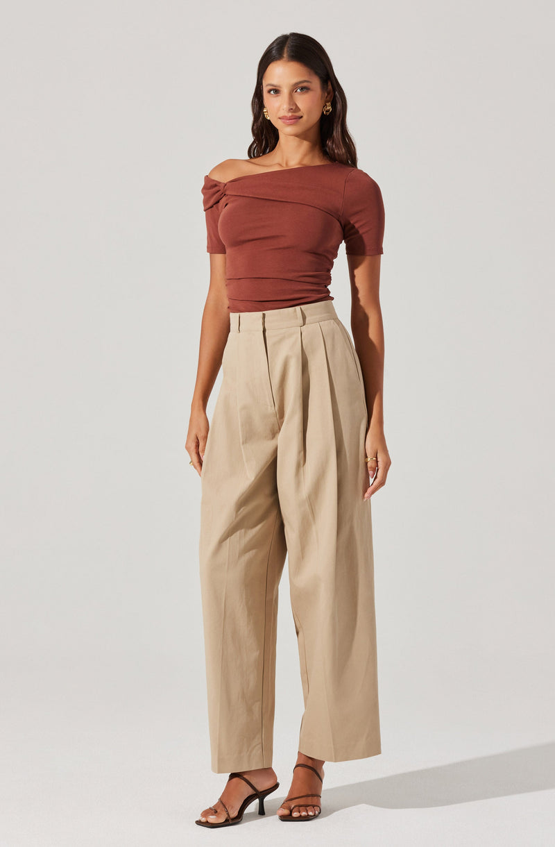 A woman is wearing a fitted, off-the-shoulder top in a rust color with horizontal pleats. She pairs it with high-waisted, wide-leg beige trousers that have pleats at the front. She completes the look with open-toe, ankle-strap high heels in a dark color.