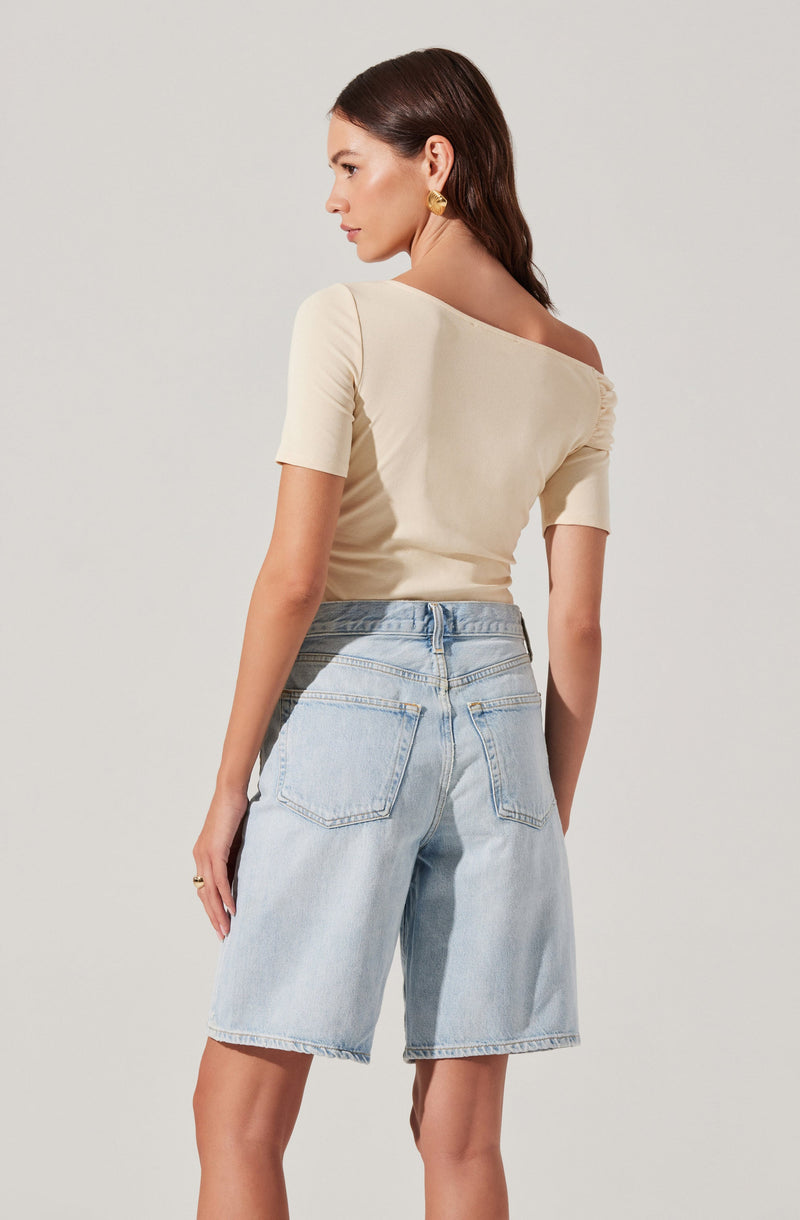 A woman is wearing a cream-colored, off-the-shoulder top with short sleeves, fitted at the waist and slightly ruched on the shoulders. She pairs it with light-wash, high-waisted denim shorts that are loose-fitting with large back pockets. The outfit has a casual and stylish appearance.