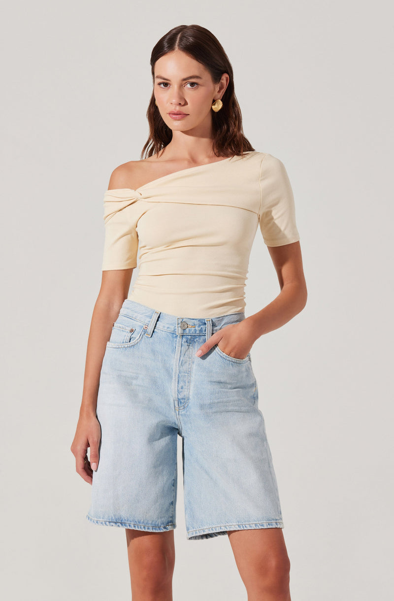 A woman is wearing a light cream-colored, off-the-shoulder top with short sleeves. The top has a twisted knot detail at the neckline and is fitted, with a ruched appearance around the waist. She is also wearing high-waisted, light-wash denim shorts that are loose-fitting and end above the knees.