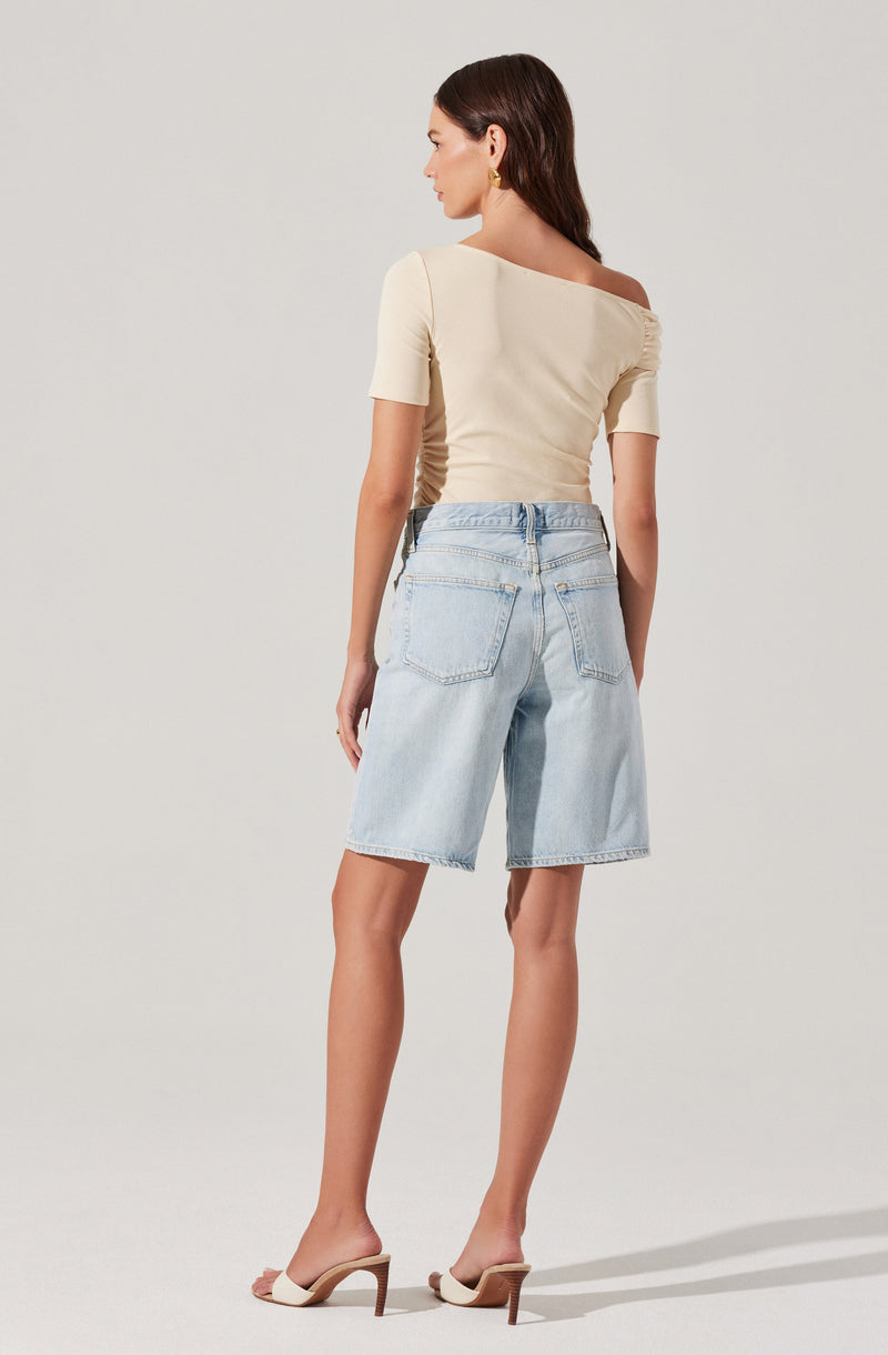 A woman is wearing a beige, off-the-shoulder top with short sleeves, paired with light blue knee-length denim shorts. She is also wearing beige high-heeled sandals with open toes and a bow detail on the front.