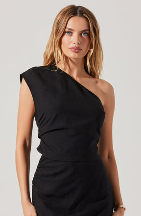 A woman is wearing a black, asymmetrical sleeveless top with a single shoulder strap on her left side, draped fabric on the right shoulder, and a fitted waist. The outfit appears elegant and stylish with a simple, sleek design.