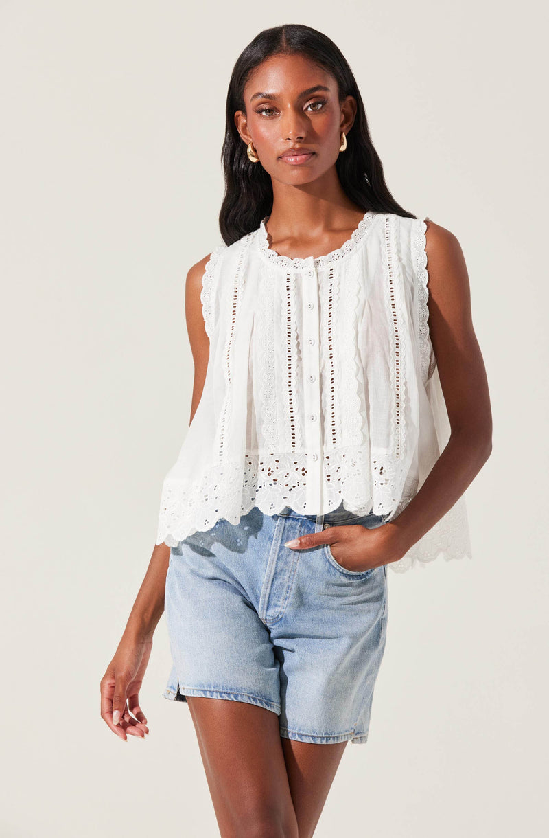 A woman is wearing a sleeveless, white embroidered top with eyelet details, scalloped edges, and a row of buttons down the front. She pairs the top with light-wash denim shorts.