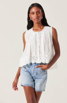 A woman is wearing a sleeveless, white embroidered top with eyelet details, scalloped edges, and a row of buttons down the front. She pairs the top with light-wash denim shorts.