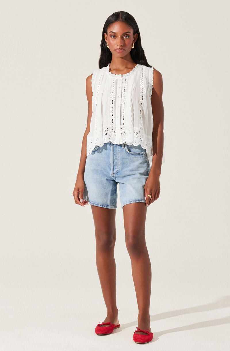 A woman is wearing a sleeveless white eyelet blouse with vertical lace and embroidered details, paired with light blue high-waisted denim shorts. She has on red ballet flats and gold hoop earrings.