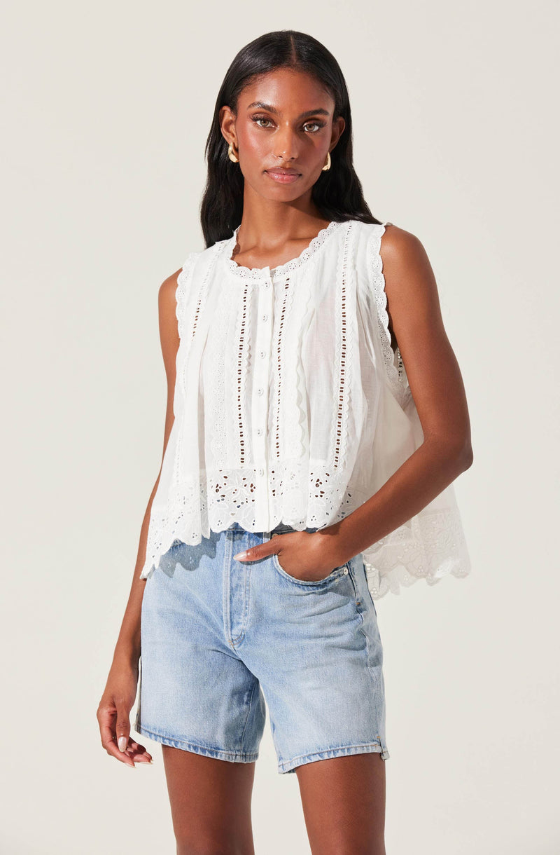 The woman is wearing a sleeveless white top with embroidered and eyelet details, featuring a scalloped hem. She pairs it with light wash denim shorts.