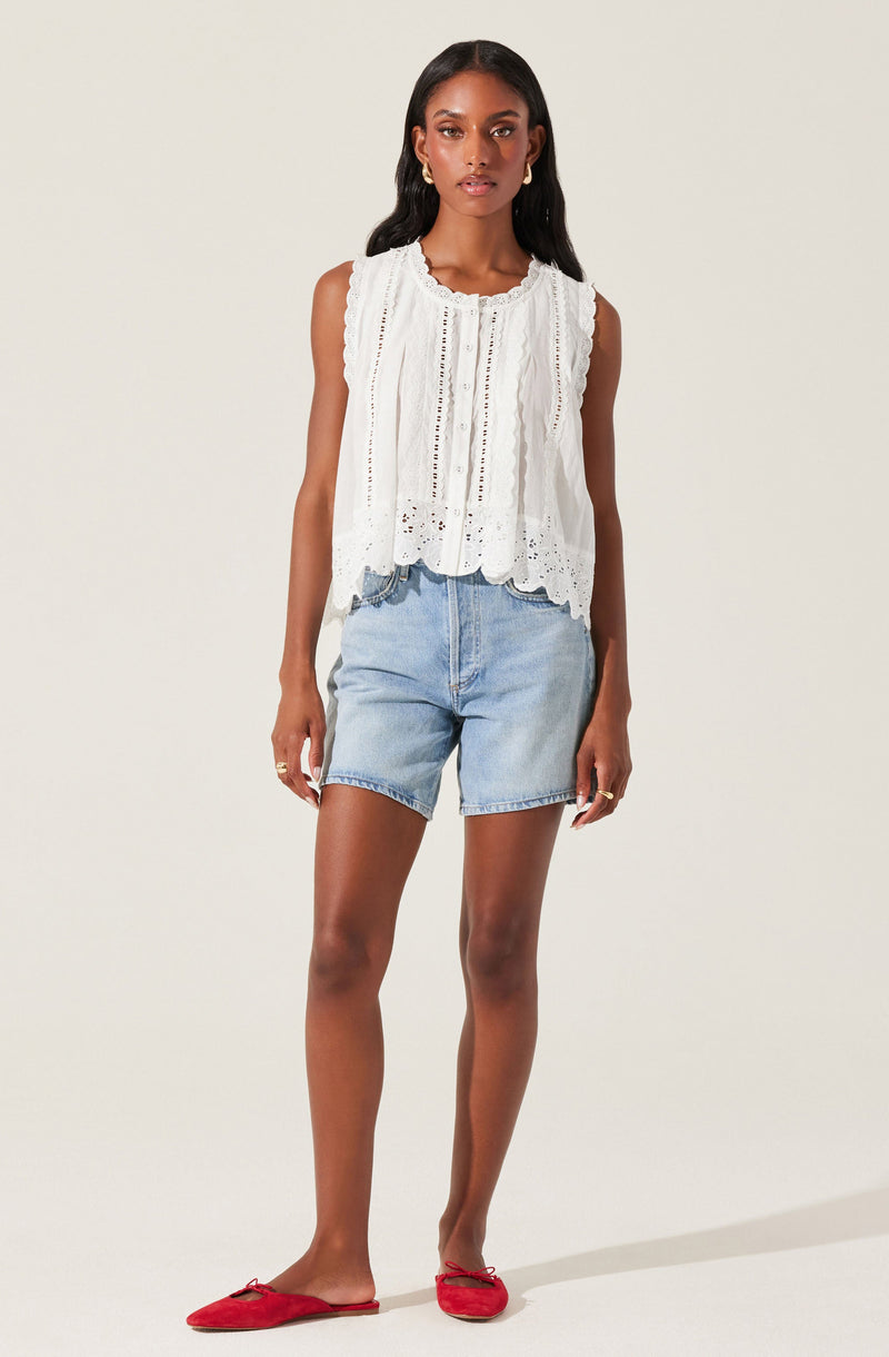 A woman wearing a sleeveless white embroidered top with eyelet patterns and scalloped edges, paired with light wash denim shorts and red flat shoes.
