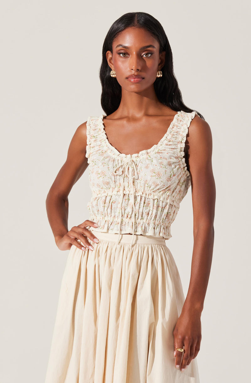 A woman is wearing a sleeveless, cropped top with ruffled shoulder straps, a gathered elastic waistband, and a delicate floral pattern in soft pastel colors. She pairs it with a high-waisted, flowing skirt in a light cream or beige color.