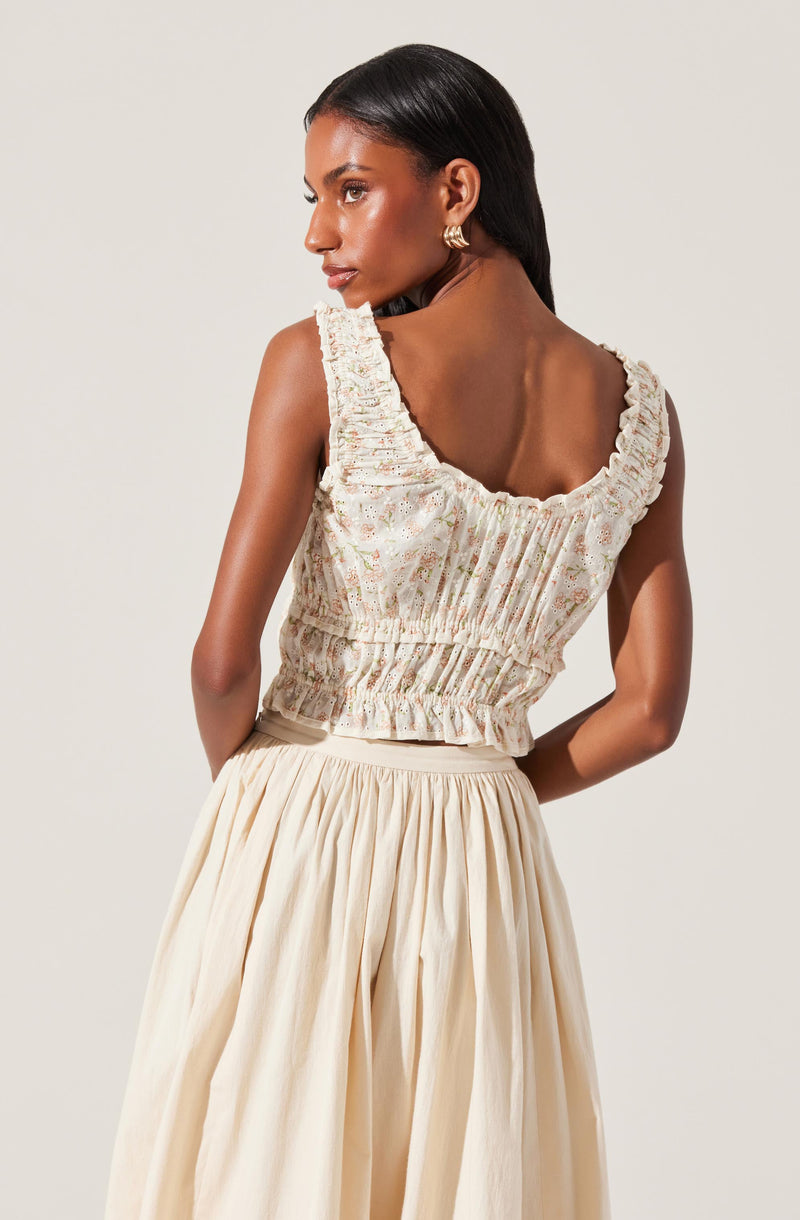 A woman is wearing a sleeveless, cropped top with ruffled shoulder straps and a gathered, elasticized waist. The top has a light, floral-patterned fabric with subtle colors. She pairs it with a high-waisted, flowing, cream-colored skirt that has pleats.