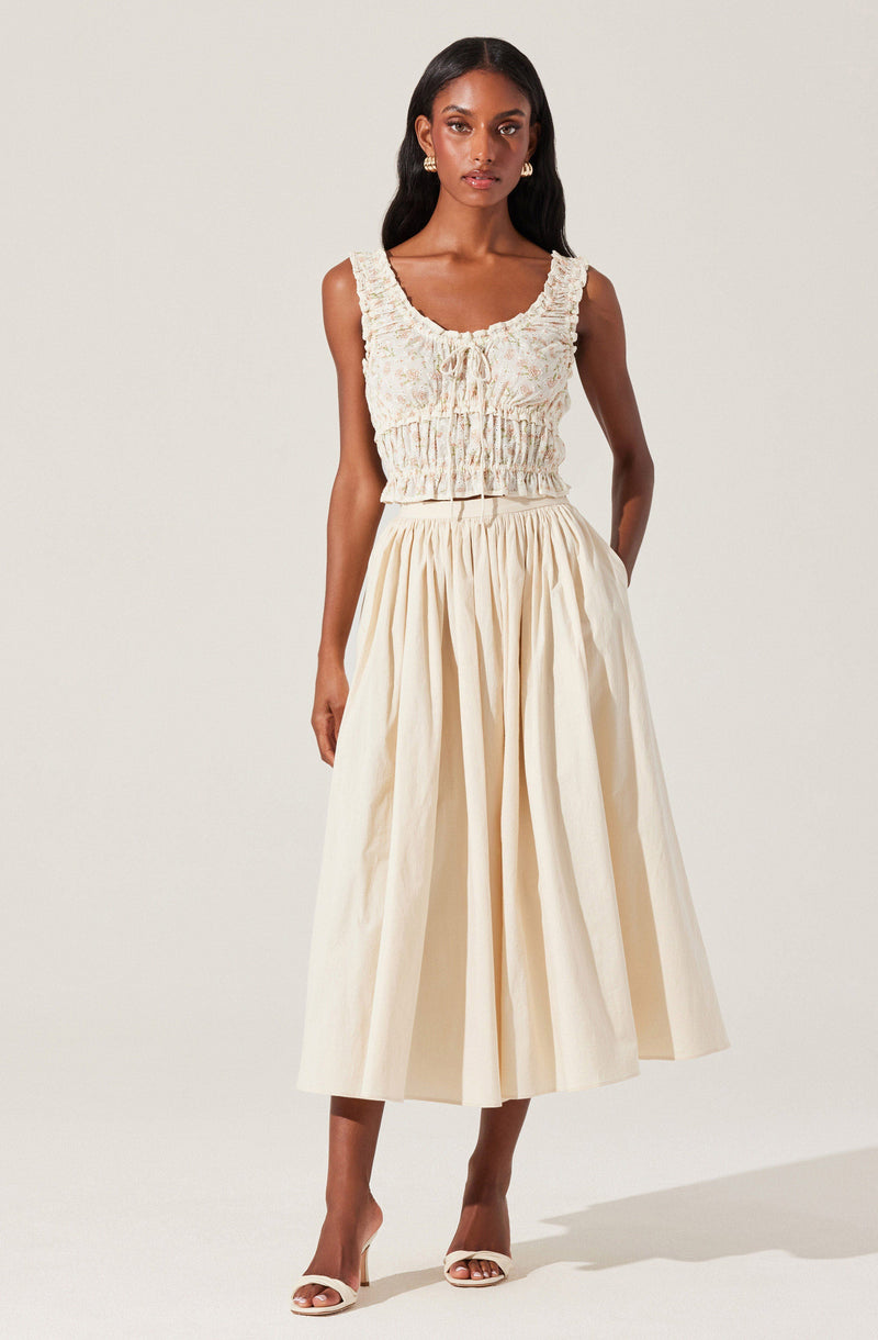 A woman is wearing a sleeveless, textured, light-colored crop top with a drawstring front and elastic waistband. She pairs it with a high-waisted, full, cream-colored midi skirt with pleats. She completes her look with open-toe heels.