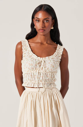 A woman is wearing a sleeveless, cropped top with ruffled shoulder straps, a gathered elastic waistband, and a front tie detail. The top features a light, pastel floral pattern. She pairs it with a high-waisted, cream-colored skirt that has a full, pleated silhouette.