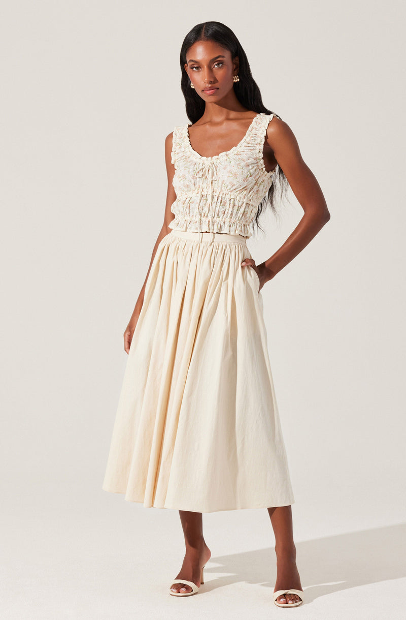 A woman is wearing a sleeveless, textured top with ruffled edges around the neckline and armholes, featuring a gathered and slightly cropped fit. She pairs it with a high-waisted, full-length, cream-colored skirt that has pleats, creating a flowing silhouette. She is wearing open-toed, heeled sandals.