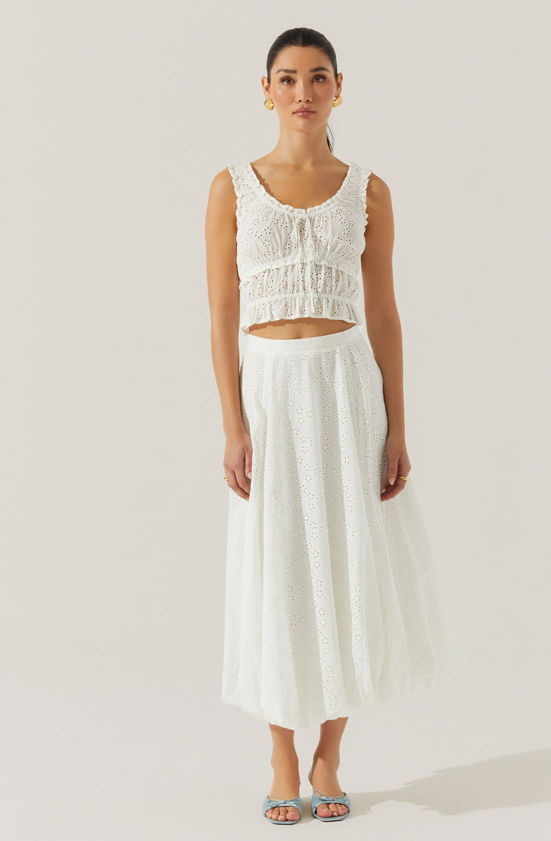 A woman is wearing a sleeveless, white, eyelet crop top with ruffled details and a scoop neckline. She pairs it with a high-waisted, white, eyelet maxi skirt that features a subtle pattern of small holes and a flowy silhouette. She completes her outfit with light-colored open-toe heels and gold earrings.