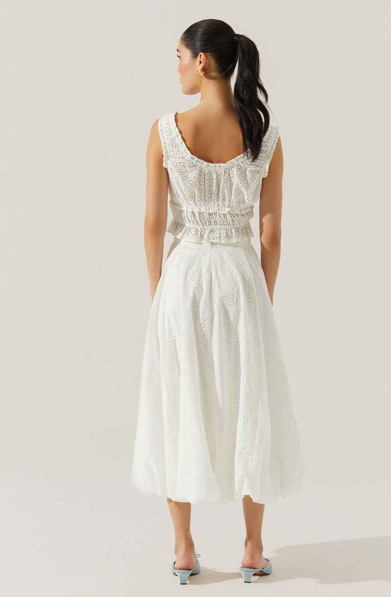 A woman is wearing a sleeveless, white eyelet dress with ruffle details on the neckline and waist. The dress has a loose, flowing skirt reaching mid-calf length. She has a high ponytail hairstyle and is wearing small hoop earrings.