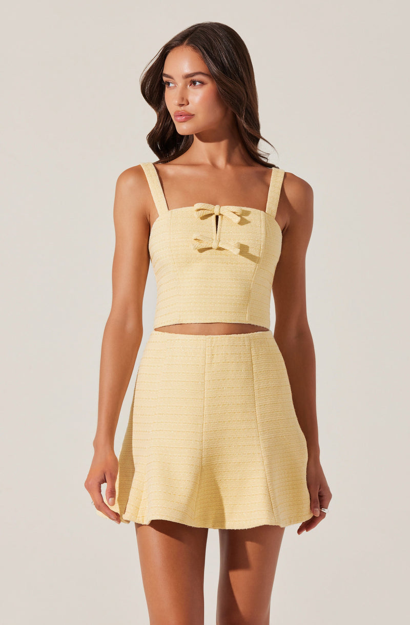 A woman is wearing a coordinated two-piece outfit in a soft yellow color. The top is a sleeveless crop top with wide straps and a decorative bow in the center of the bust area. The high-waisted skirt has a textured fabric with a horizontal stripe pattern and a slight flare at the hem.