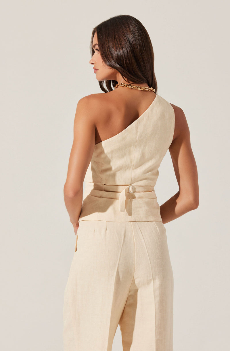A woman is wearing a sleeveless, cream-colored outfit with a one-shoulder top that has a fitted waist, secured with a matching fabric belt. She is also wearing matching high-waisted trousers with a loose fit. She accessorizes with a chunky gold chain necklace.