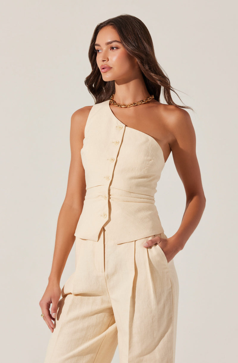 A woman is wearing a sleeveless, cream-colored, button-up top with an asymmetrical design that covers one shoulder while leaving the other shoulder bare. She pairs it with matching high-waisted, wide-leg trousers that have a relaxed fit. The outfit appears to be made of a lightweight, textured fabric. She accessorizes with a chunky gold chain necklace.