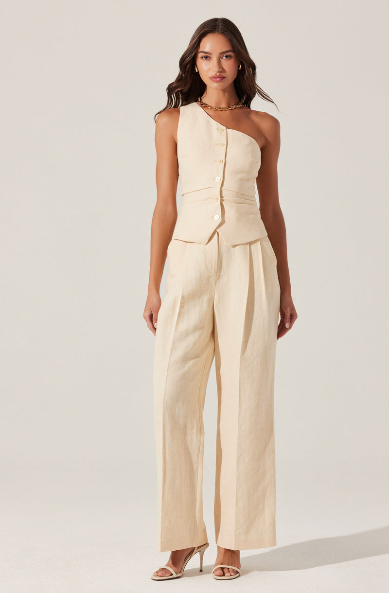 A woman is wearing a sleeveless, asymmetrical cream-colored top with a button-down front and matching high-waisted, wide-leg pants. She accessorizes with gold jewelry, including hoop earrings and a chain necklace, and is wearing open-toe high heels.