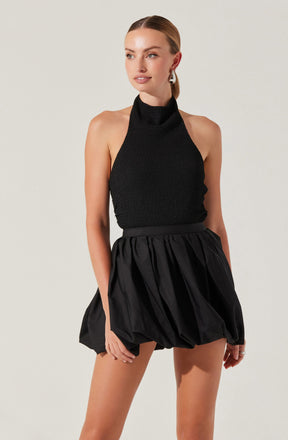 A woman wearing a sleeveless black textured halter top with a high neckline, paired with a high-waisted black skirt that has a voluminous, bubble style with gathered fabric. She has her hair pulled back and is accessorized with signature earrings.