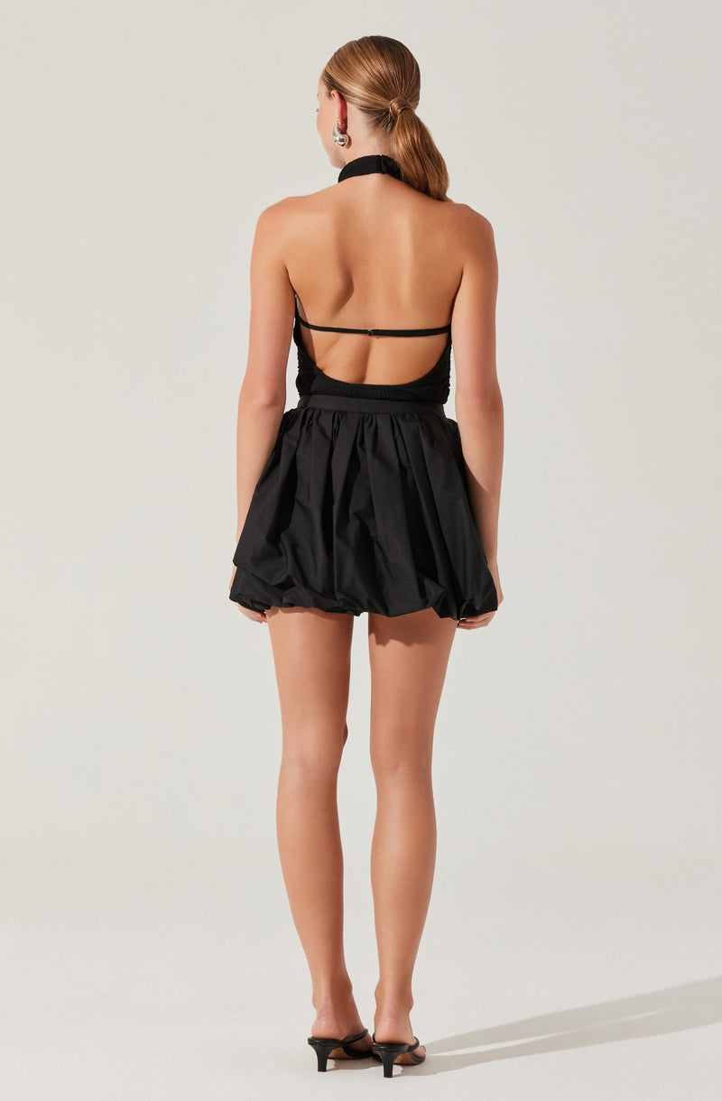 A woman is wearing a backless black top with thin straps and a high neckline, paired with a voluminous black skirt that has a gathered, puffy design. She is also wearing black heeled sandals and has her hair styled in a low ponytail.