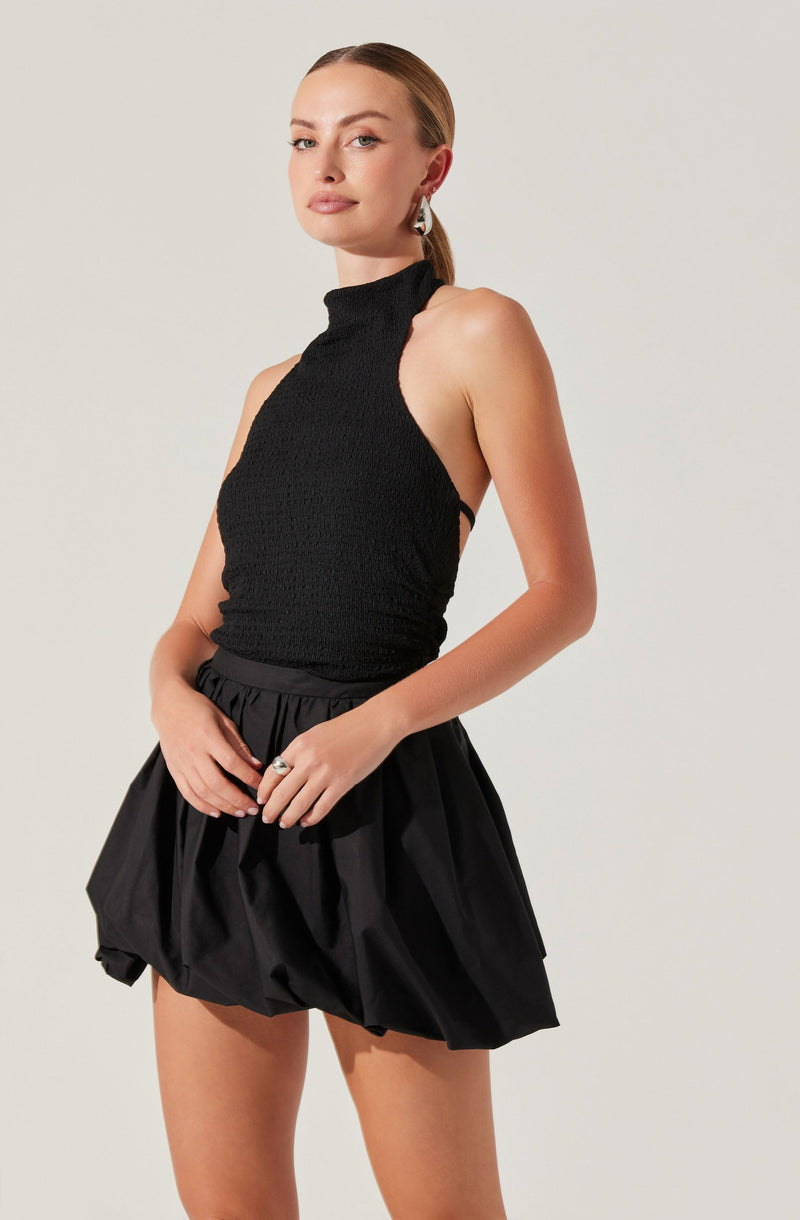 A woman is wearing a sleeveless black top with a high neckline and textured fabric. She is also dressed in a black, voluminous, bubble-style skirt with gathered details. She accessorizes with large, silver hoop earrings and a ring on her right hand.