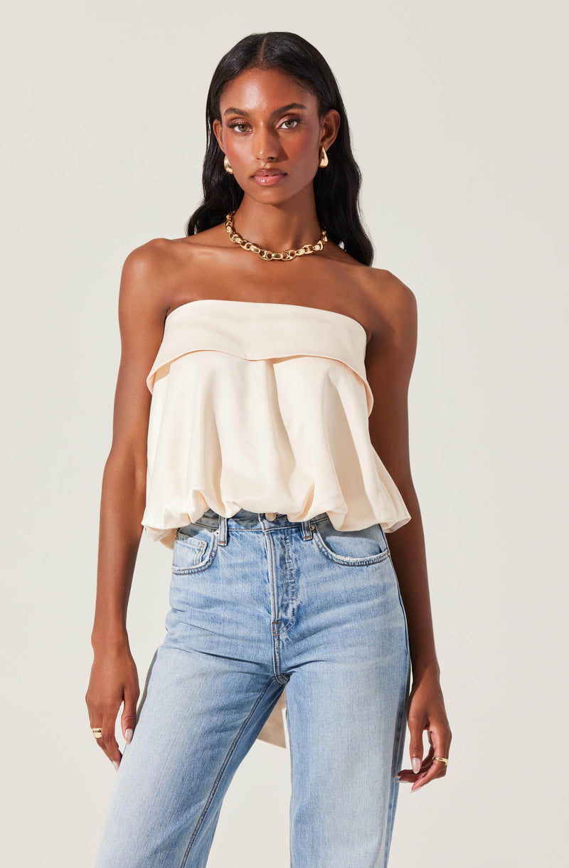 A woman is wearing a strapless, off-white, ruffled top with a loose, flowing fit. She has paired it with high-waisted light blue jeans. She accessorized with a chunky gold chain necklace, large gold hoop earrings, and a gold ring on her right hand.