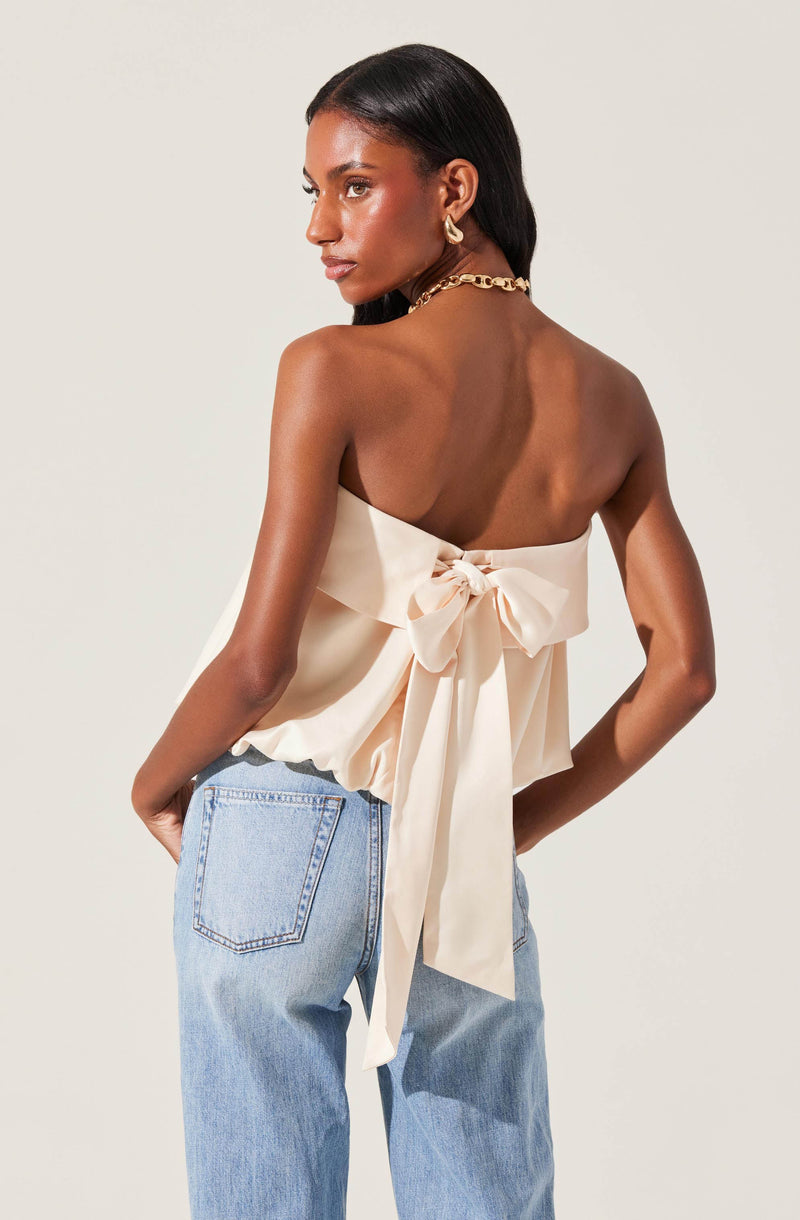 A woman is wearing a sleeveless, off-white satin top with a large bow tied at the back, creating a ruffled effect around the waist. She pairs this with light blue, high-waisted jeans. She accessorizes with gold hoop earrings and a chunky gold chain necklace.