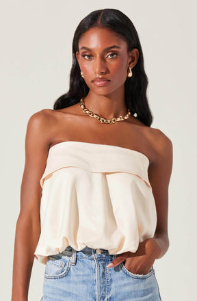 A woman is wearing a strapless, cream-colored, loose-fitting top with a wide band around the top edge. The top has a gathered, billowing design around the waist. She is also wearing a chunky gold chain necklace and gold hoop earrings. The outfit is paired with light-wash denim jeans.