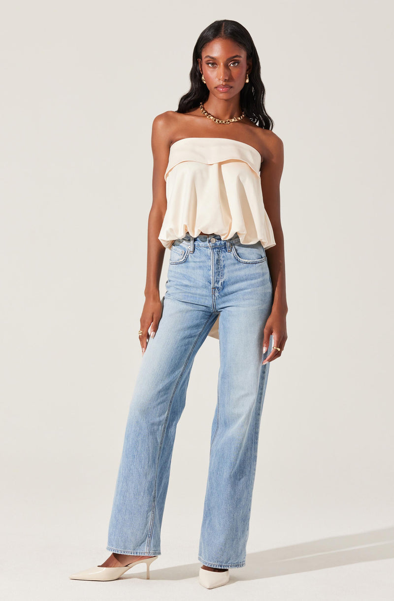A woman wearing a strapless, off-white, ruffled top that is loose and billowy, paired with high-waisted light blue jeans and white pointed-toe heels. She accessorizes with a gold necklace and earrings.