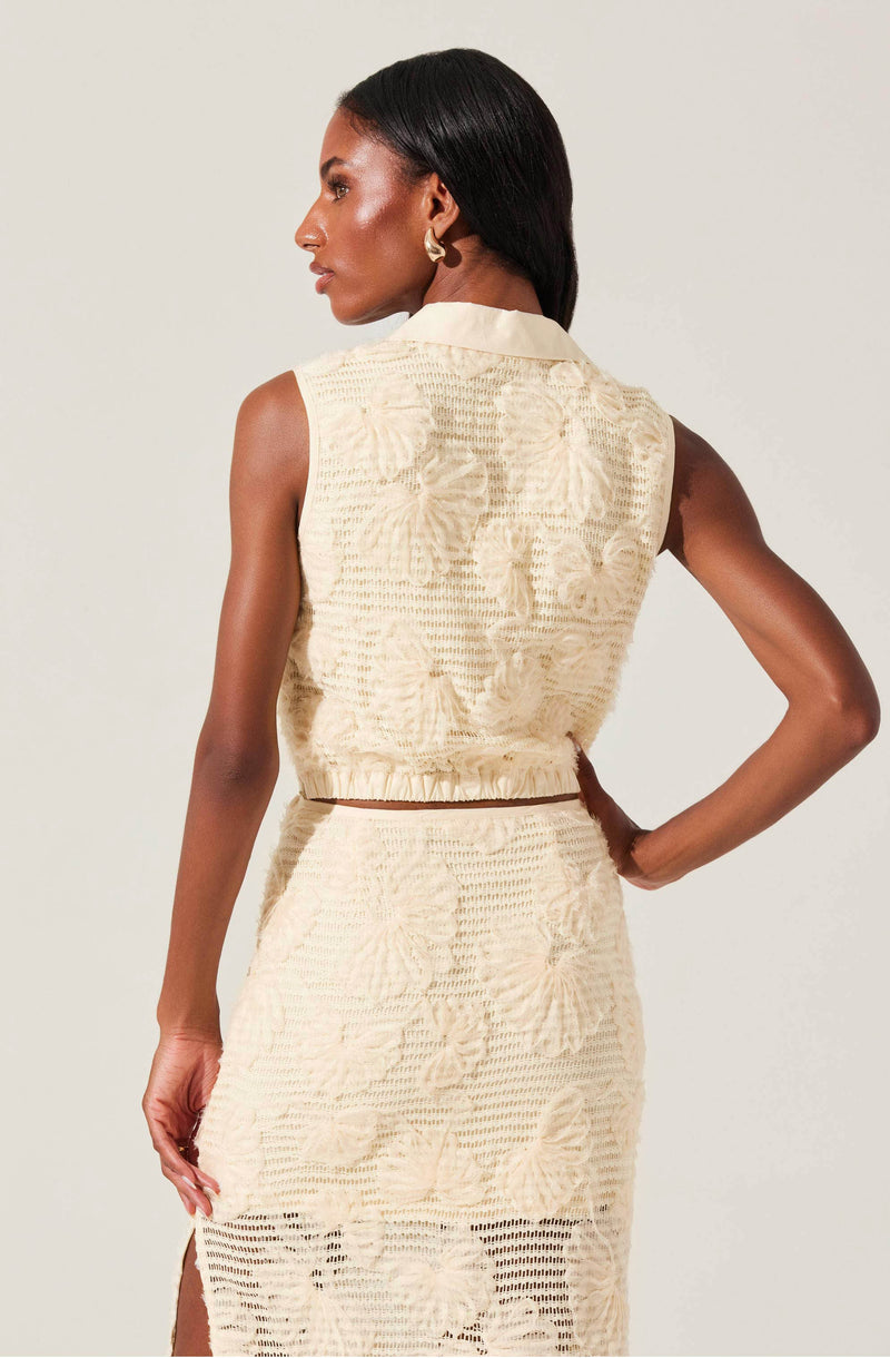 A woman with dark hair styled straight is wearing a sleeveless, cream-colored textured dress with a floral pattern. The top has a collar, and the bottom is a matching skirt with a slit on the side, both made from a semi-sheer fabric with intricate embroidery.